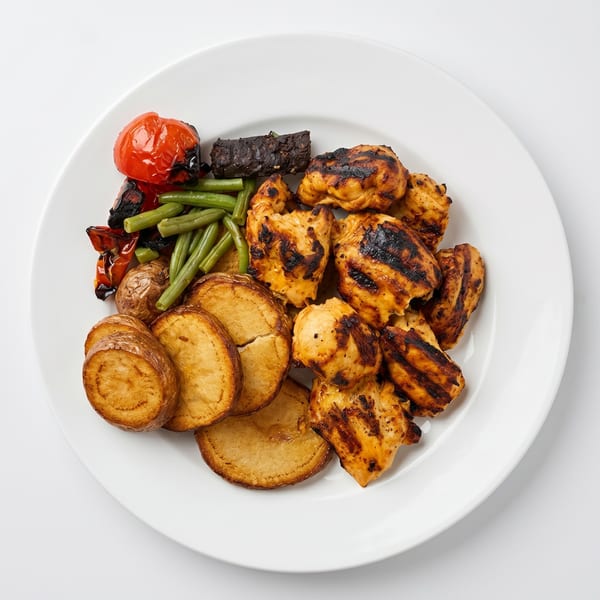 Grilled chicken pieces, roasted potatoes, green beans, and charred tomatoes on a white plate.